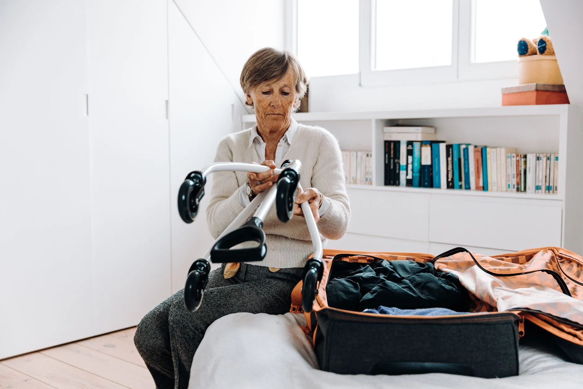 Senior woman preparing for travel, packing suitcase with walker in bright modern bedroom.