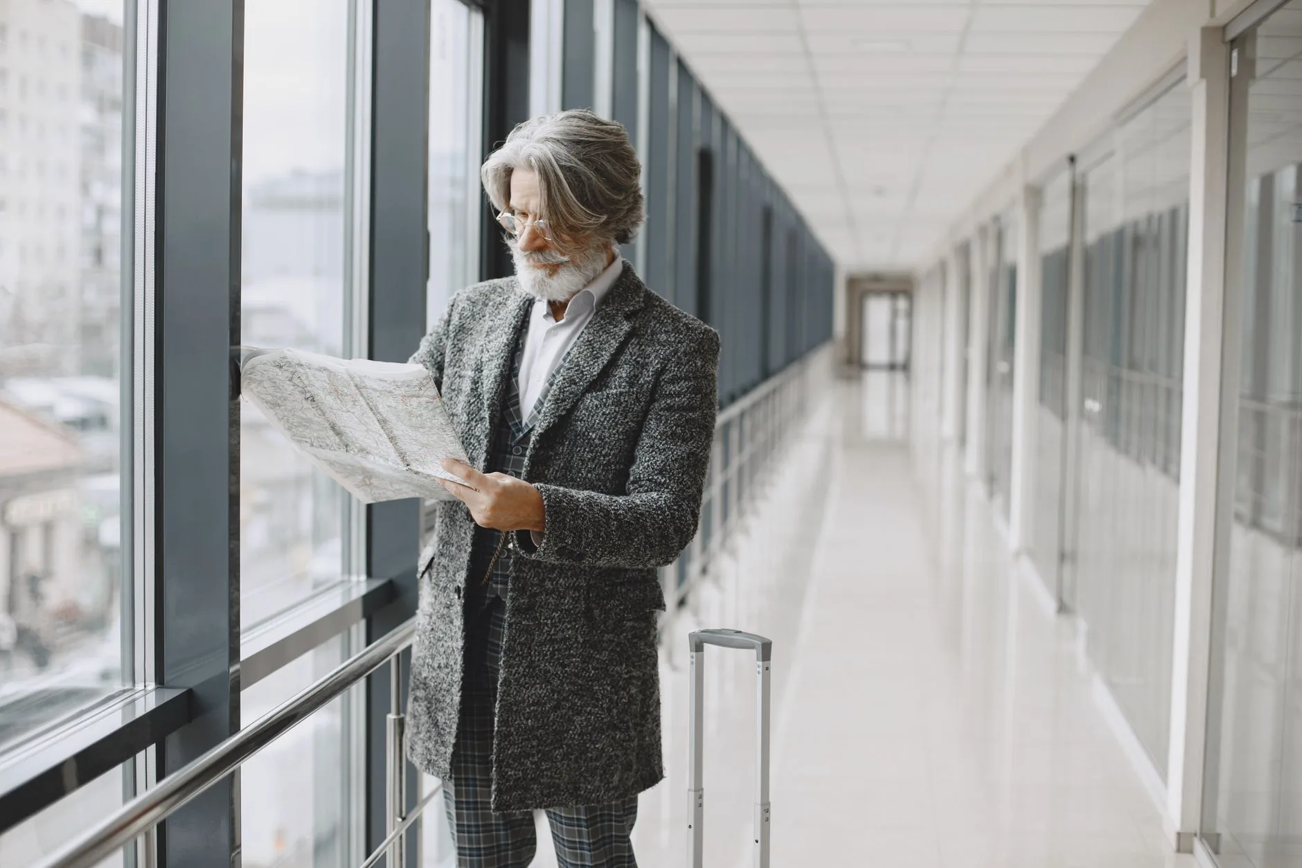 Senior man with beard reads map by glass windows in modern hallway with luggage.